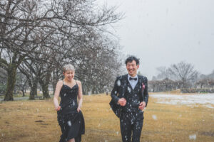 雪の中で富山県中央植物園の冬の洋装ロケーションフォトウェディングで、雪の中走って笑い合う黒ドレスと黒スーツの新郎新婦の楽しそうなシーン。