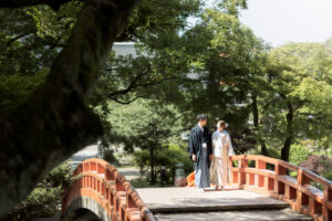 富山の城址公園で和装ロケーションフォトウェディングをする新郎新婦、赤い橋の上を見つめ合いがなら歩いている