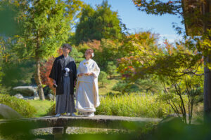 富山城址公園の日本庭園で、白無垢と紋付袴の和装を着た新郎新婦が池の上の石橋でカメラ目線で微笑んでいるフォトウェディングのシーン。