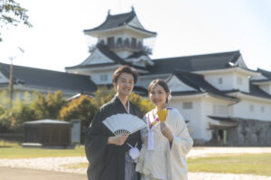 富山の城址公園で和装ロケーションフォトウェディングをする新郎新婦、白の前で扇子を広げて笑顔で写真を撮る様子